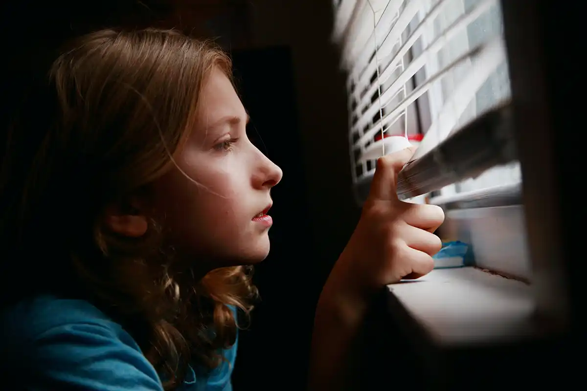 Tween boy with long hair looks out window through blinds.