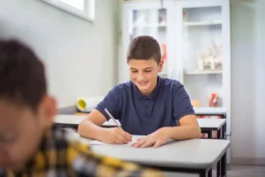 Middle school aged boy sits at desk in class working on an assignment and smiling.
