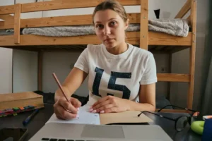 Female college student looks at camera with a vague smile as she sits at a desk in her dorm room holding a pen poised to write in a notebook.